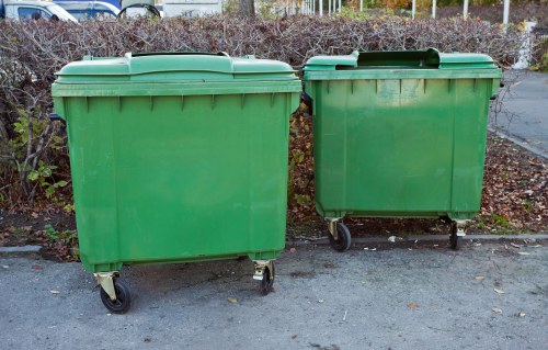 Two large green plastic commercial wheelie bins with black wheels and lids, positioned side by side on a paved surface in an outdoor setting. The bins are situated in front of shrubbery and low hedges, with a background of a paved area and a fence. The left bin is slightly larger and taller, while the right bin appears more compact. Both bins are used for rubbish collection and disposal, consistent with waste management services in Carshalton. The scene is illuminated by natural daylight, providing clear visibility of the textured surface of the bins and surrounding environment, relevant for a garden clearance or rubbish removal service in the local area operated by Garden Clearance Carshalton.