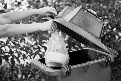 A pair of hands placing a plastic bag filled with white crockery into the open lid of a large, foot-operated black wheelie rubbish bin situated outdoors, with a background of dense leafy bushes suggesting a garden or backyard setting. The bin has a textured surface and a sturdy lid, positioned on a paved or concrete surface. The scene reflects regular rubbish disposal activities and aligns with services provided by Garden Clearance Carshalton for waste and rubbish removal, particularly in residential areas within the local postcode area. The natural lighting indicates daytime, with shadows cast by the lid and hands. The focus is on the practical aspect of rubbish handling, with the plain black bin and the clear, covered contents demonstrating typical domestic waste management practices relevant to garden or household clearance tasks in the Carshalton area.