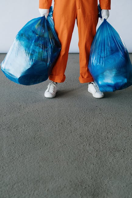 A person wearing a white shirt and beige trousers holds a transparent plastic bottle with a red cap above a recycling bin labeled 'PLASTIC.' The individual's red cardigan is partially visible on their